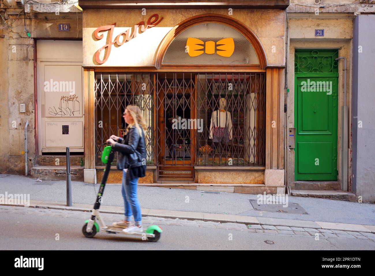 Una donna su uno scooter elettrico a noleggio (trottinettes électriques) zips passato le Colonel Moutarde cravatta negozio su Rue Romarin, Lione, Francia Foto Stock