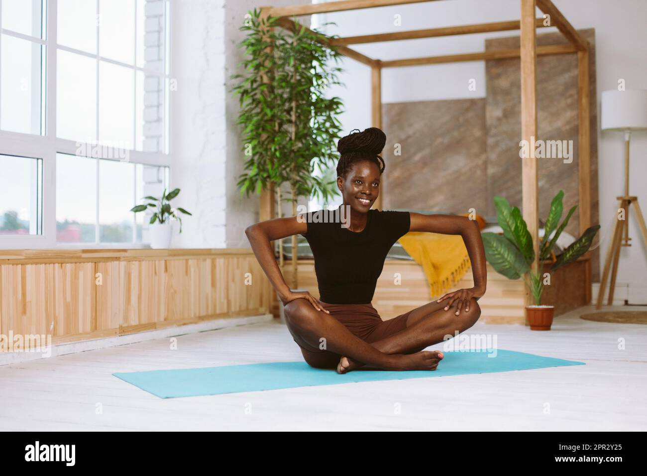Bella e atletica afro donna americana con sorriso seduta in posa farfalla, facendo yoga sul tappetino ginnico in studio decorato. Stretching muscolare Foto Stock