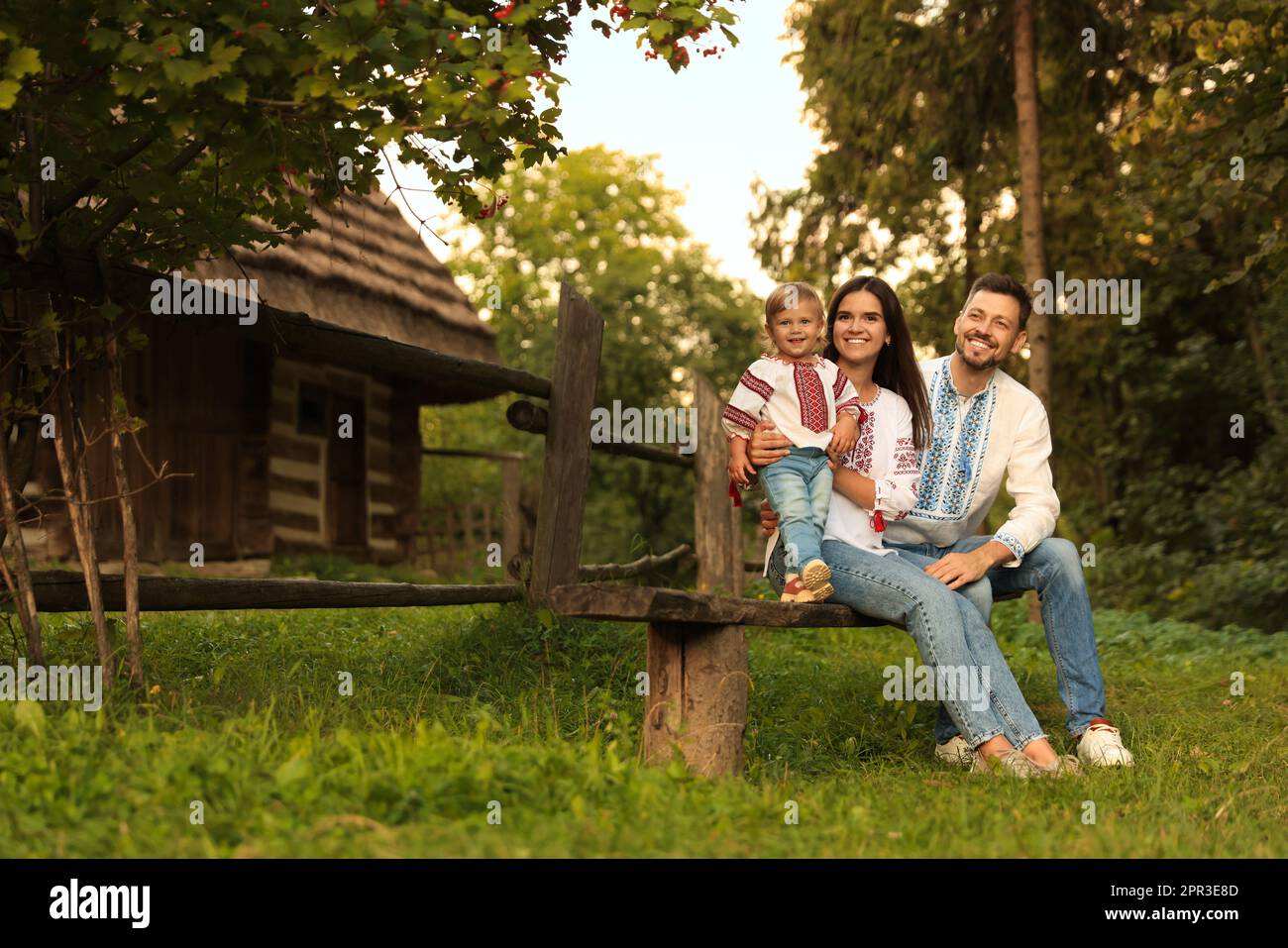 Felice cute famiglia in camicie ucraine ricamate seduto su rustico panca di legno vicino a casa. Spazio per il testo Foto Stock