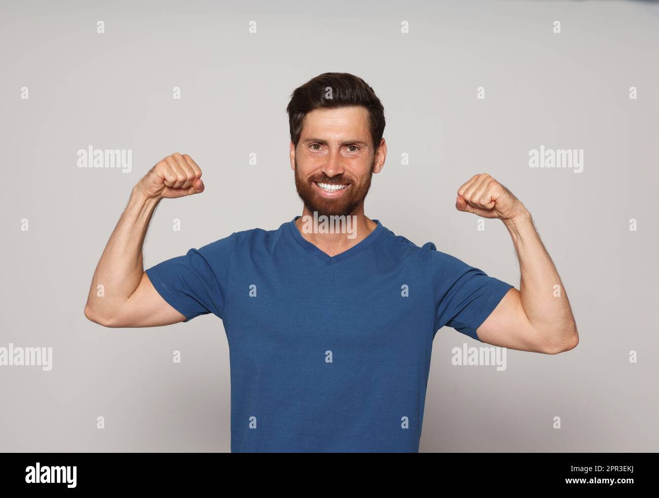 Uomo sorridente barbuto che mostra bicipiti su sfondo grigio Foto Stock