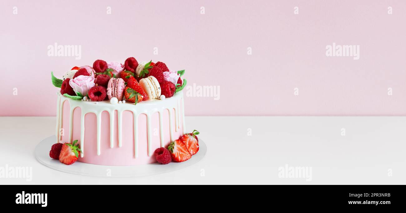 Panorama di dolce di compleanno con bacche, macaron e decorazioni floreali sul tavolo. Bella torta rosa decorata con macaron, lamponi, fragole Foto Stock