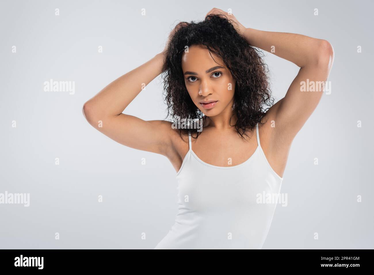 giovane afroamericana donna in bianco canotta top toccare capelli brunette ondulati e guardare la macchina fotografica isolato su grigio, immagine stock Foto Stock