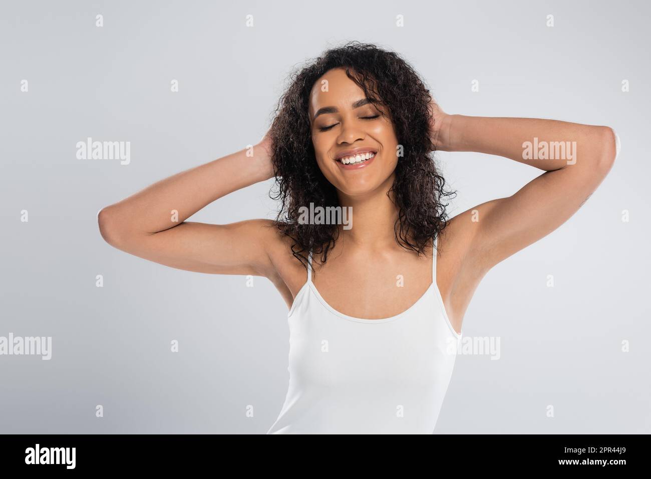 donna afro-americana spensierata in canotta bianca che tocca i capelli ondulati e sorridente con gli occhi chiusi isolato sul grigio, immagine stock Foto Stock