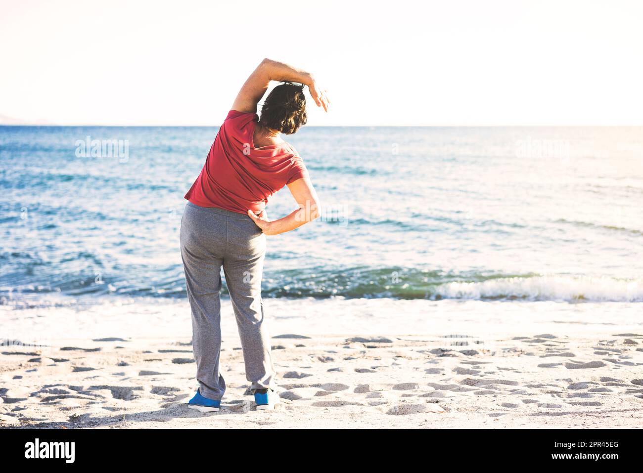 Donna anziana che fa l'esercitazione alla spiaggia. Stretching laterale, ripresa dalla parte posteriore, persona irriconoscibile. Foto Stock