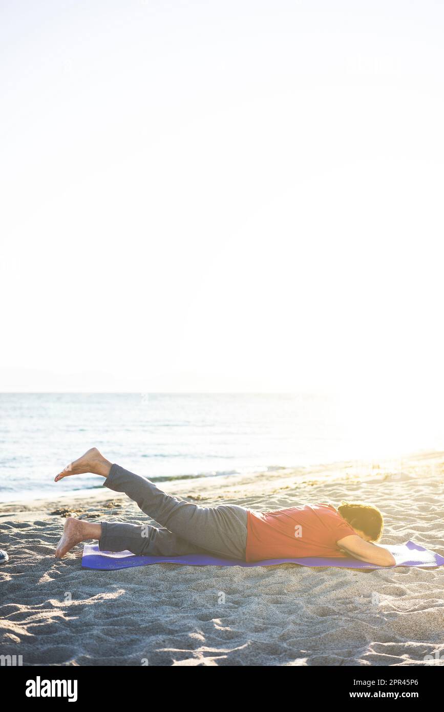 Donna anziana in spiaggia, facendo esercizio sul tappetino yoga. Invertire i sollevamenti delle gambe. Foto Stock