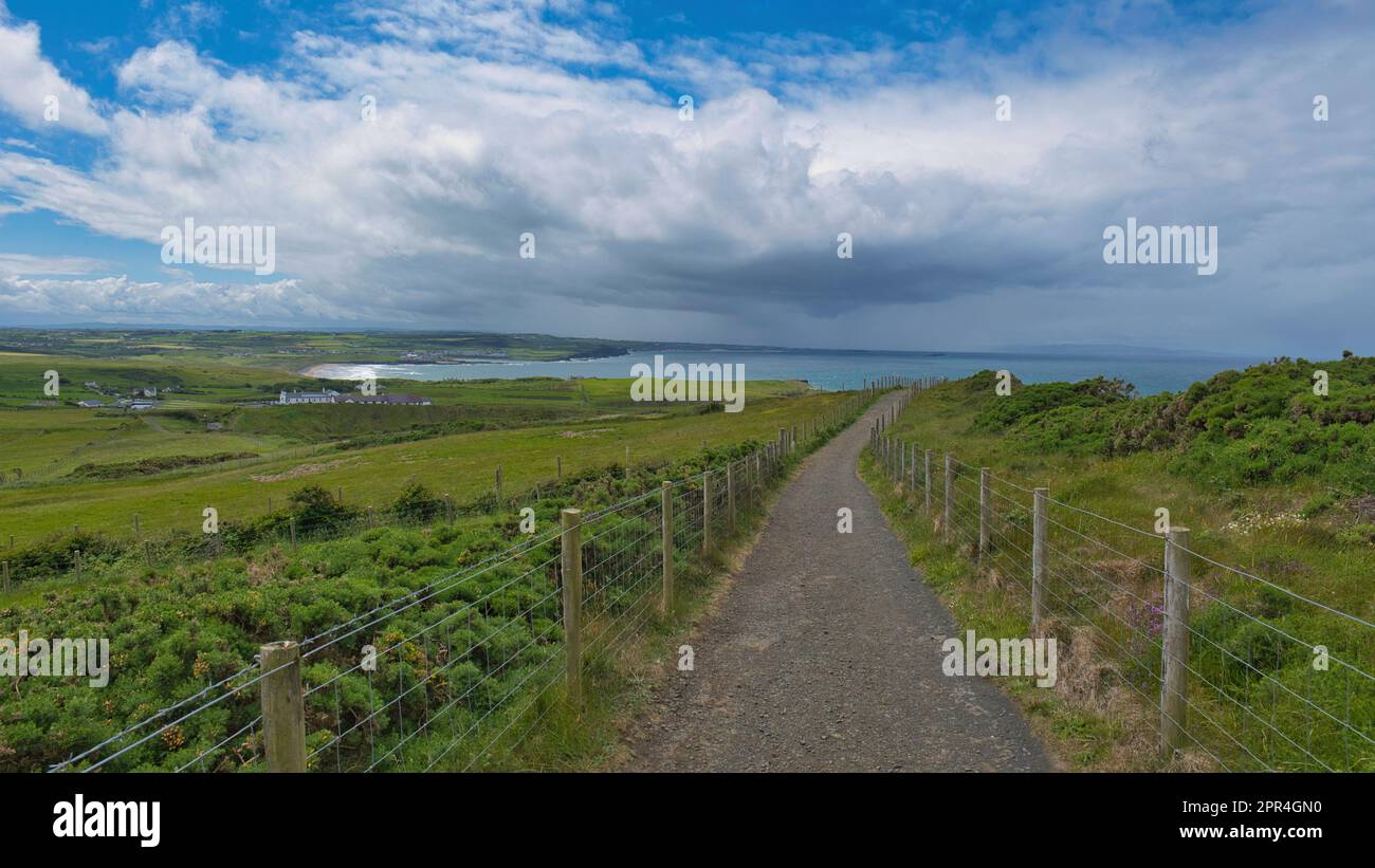 Sentieri per passeggiate ed escursioni sulle verdi colline del Selciato del gigante, Irlanda, con vista sull'oceano, cielo blu con nuvole e un villaggio irlandese sullo sfondo Foto Stock