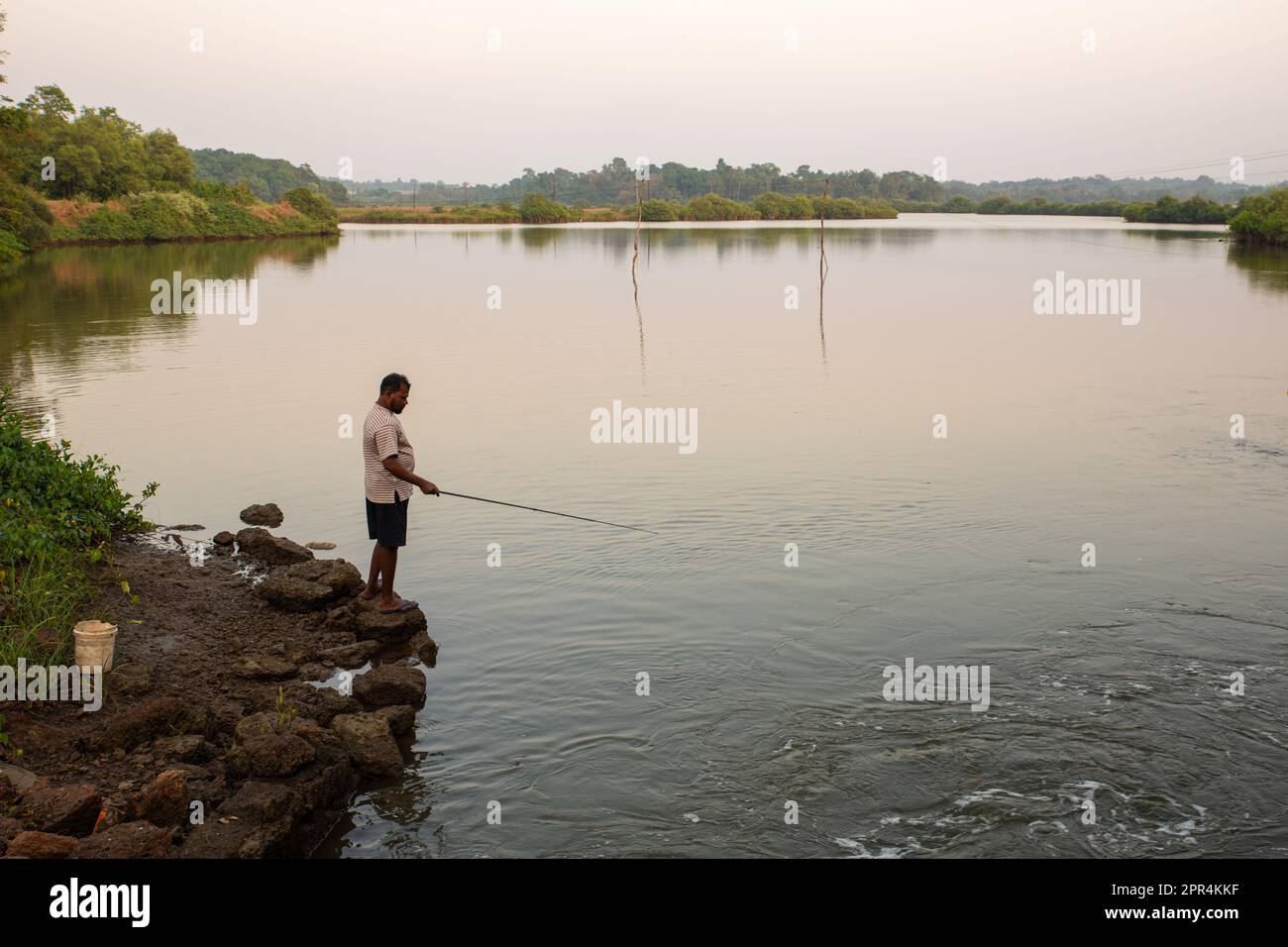 Goa, India - 19th dicembre, 2022 : un pescatore solista che si trova sulle rive di un fiume e che utilizza una canna da pesca per catturare i pesci in tarda serata. Foto Stock