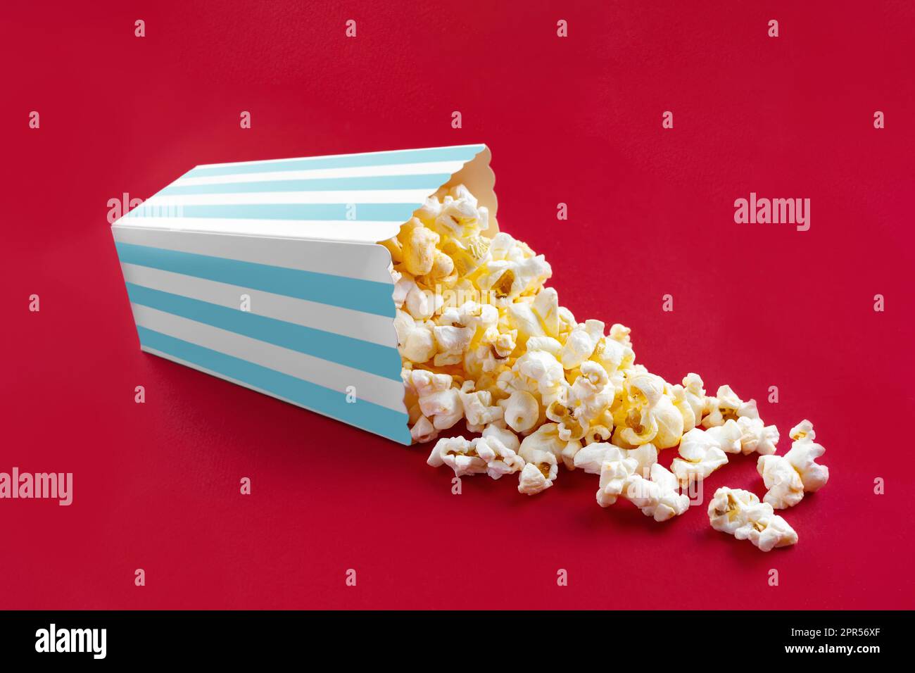 Gustoso popcorn al formaggio che cade da un secchio di cartone a righe turchesi, isolato su sfondo rosso. Dispersione di grani di popcorn. Film, cinema e it Foto Stock