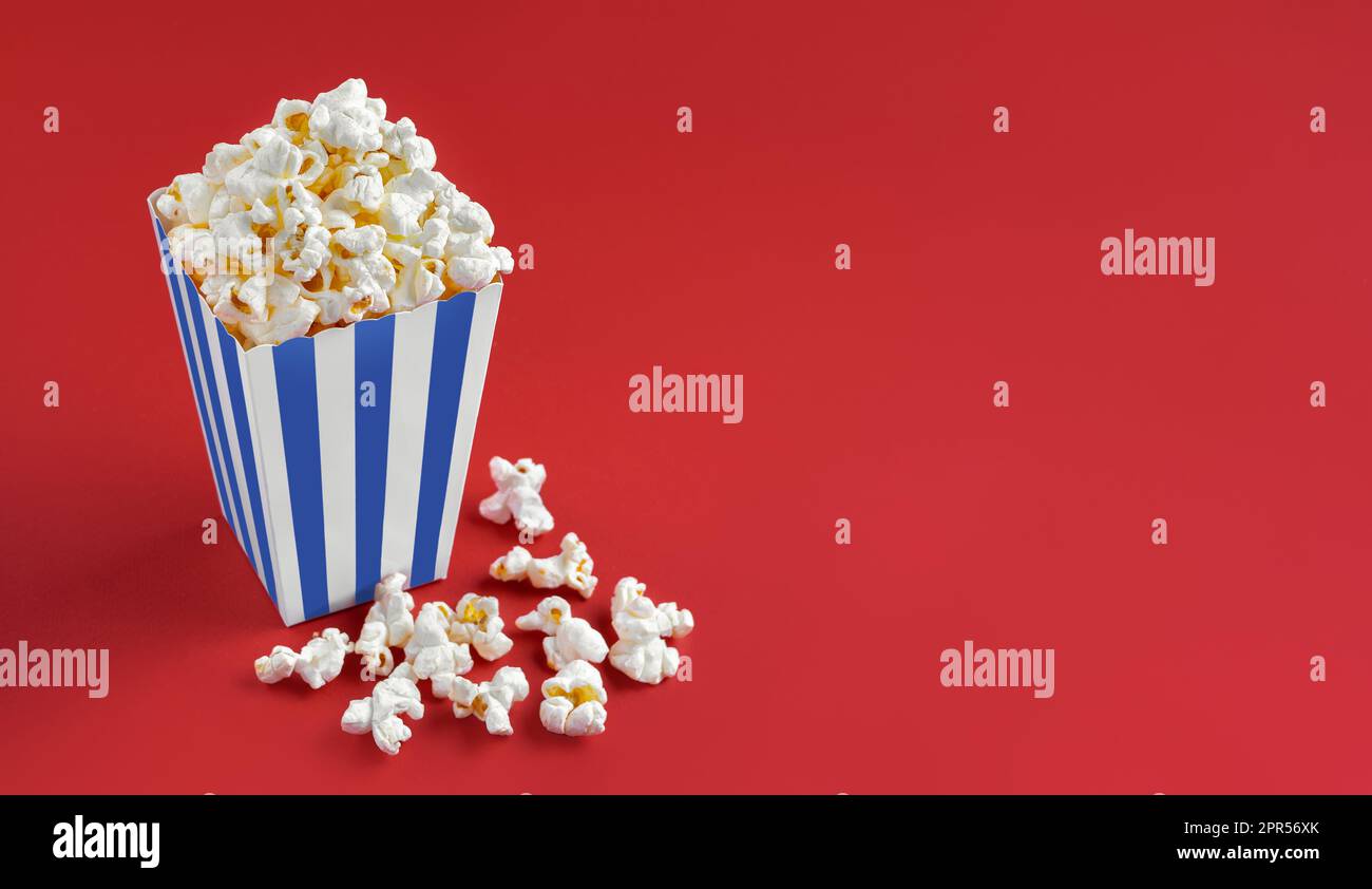 Secchio di cartone a strisce bianche blu con gustoso popcorn al formaggio, isolato su sfondo rosso. Scatola con dispersione di grani di popcorn. Fast food, film, cine Foto Stock