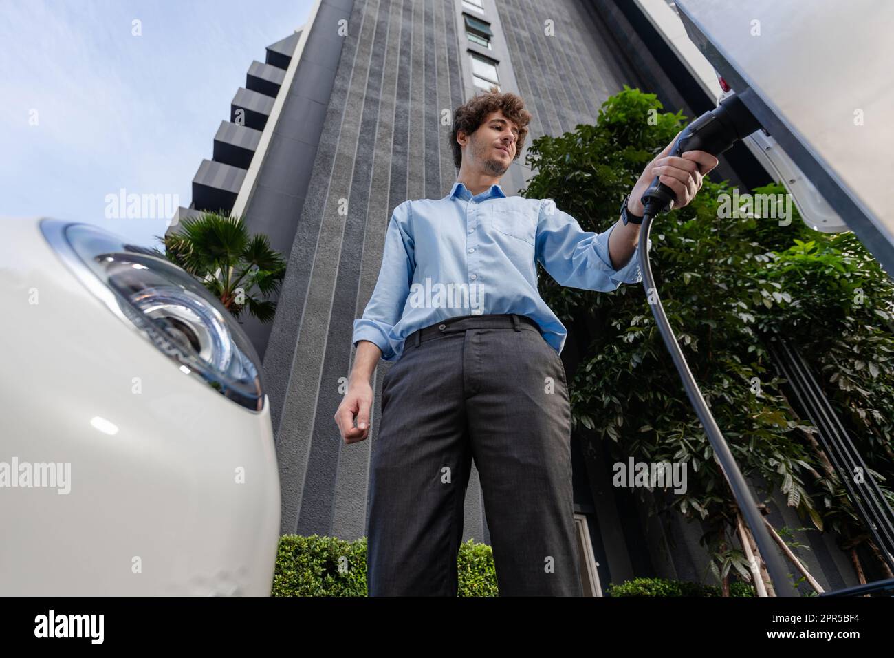 Fisheye vista vestito-rivestito uomo d'affari progressivo alla stazione di ricarica per veicolo elettrico nella città moderna con l'edificio residenziale condominio appartamenti Foto Stock