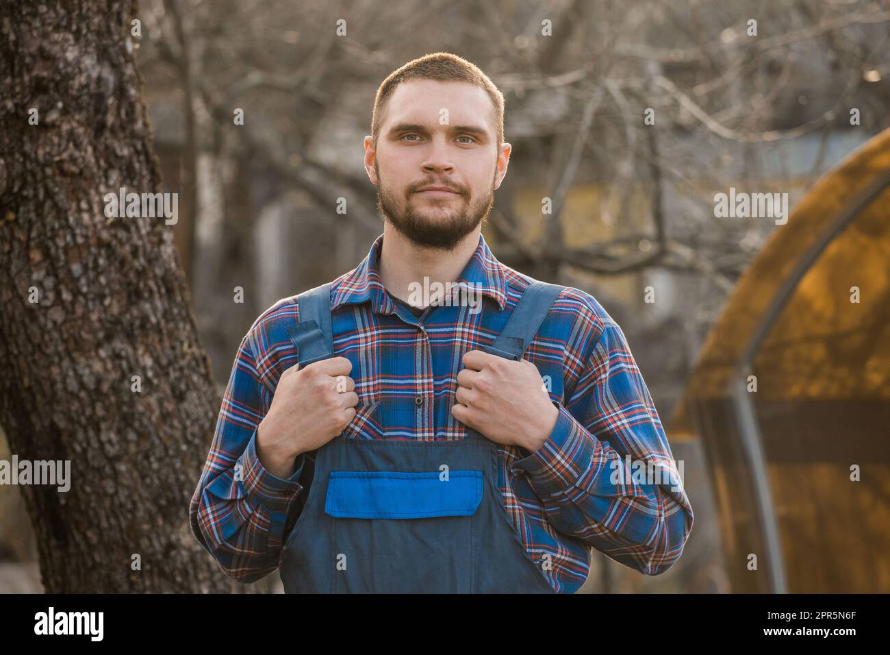 Farmer soddisfatto europeo aspetto maschile ritratto rurale con barba, camicia e tute uomo guardando la fotocamera all'aperto. Foto Stock
