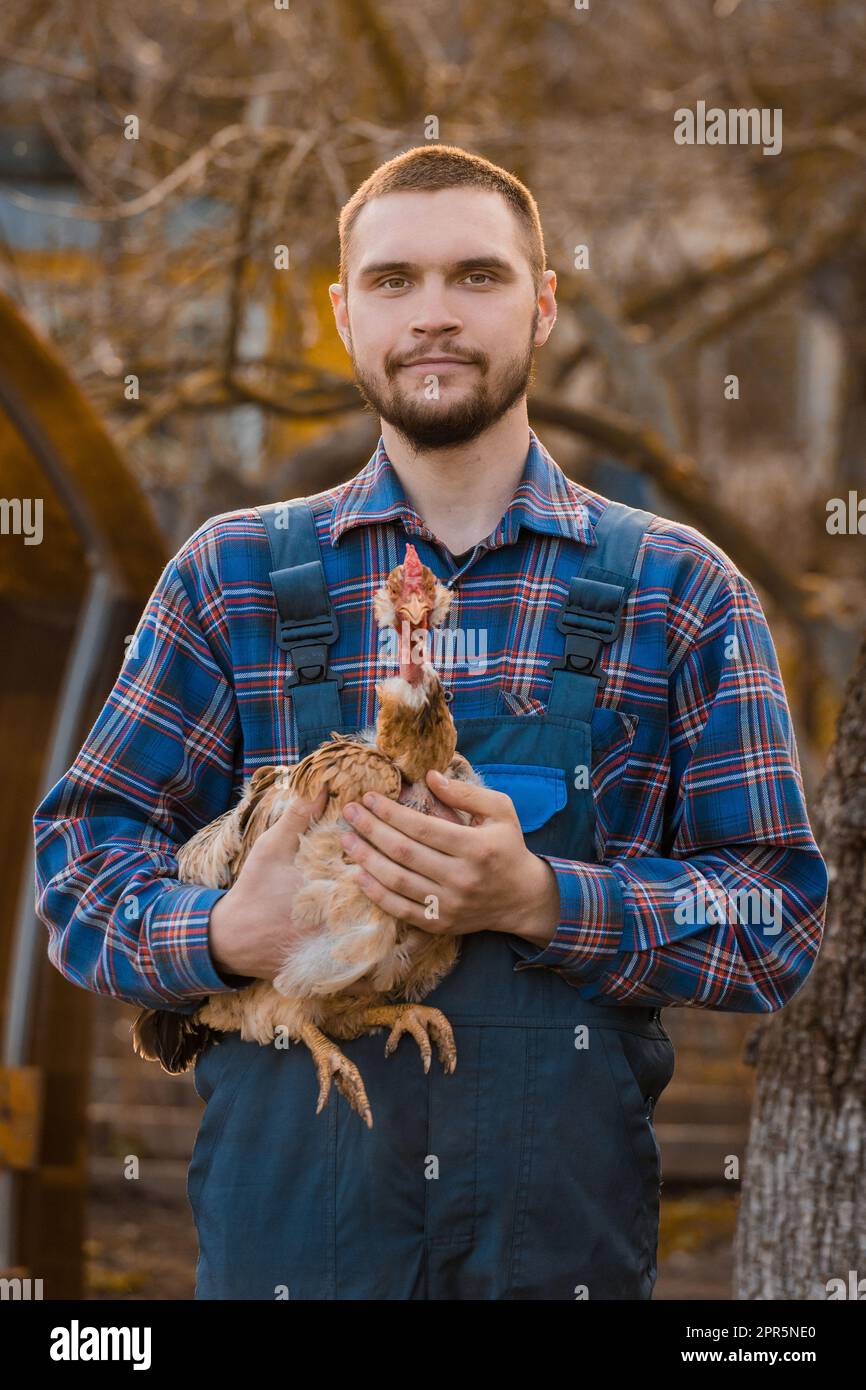 Ritratto rurale maschile con barba, camicia e tute con un pollo a collo bianco in braccio all'aperto. Foto Stock