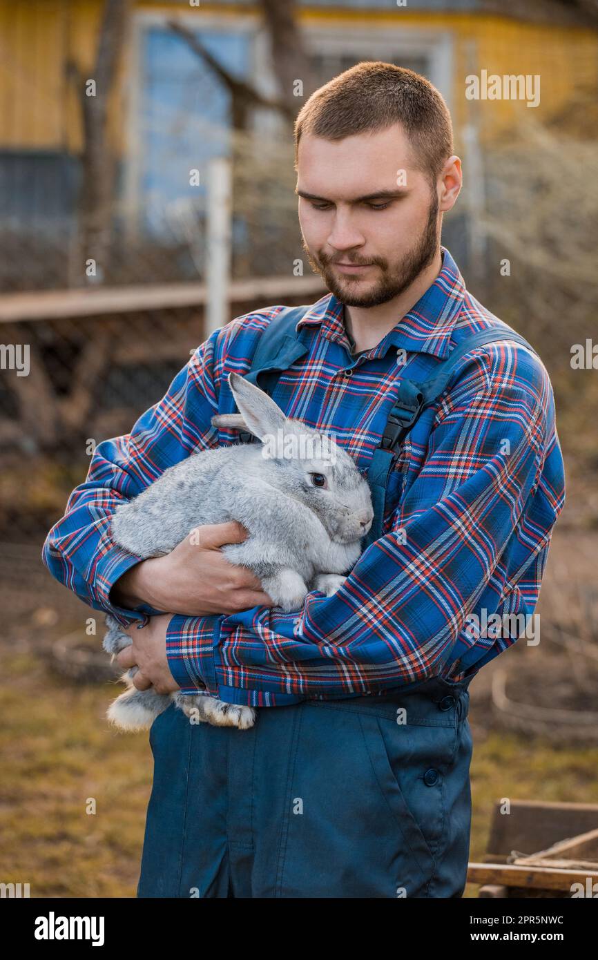 Contadino bel ritratto rurale caucasico europeo sorridente in campagna con barba, camicia e tute guarda un coniglio bianco tra le braccia su t Foto Stock