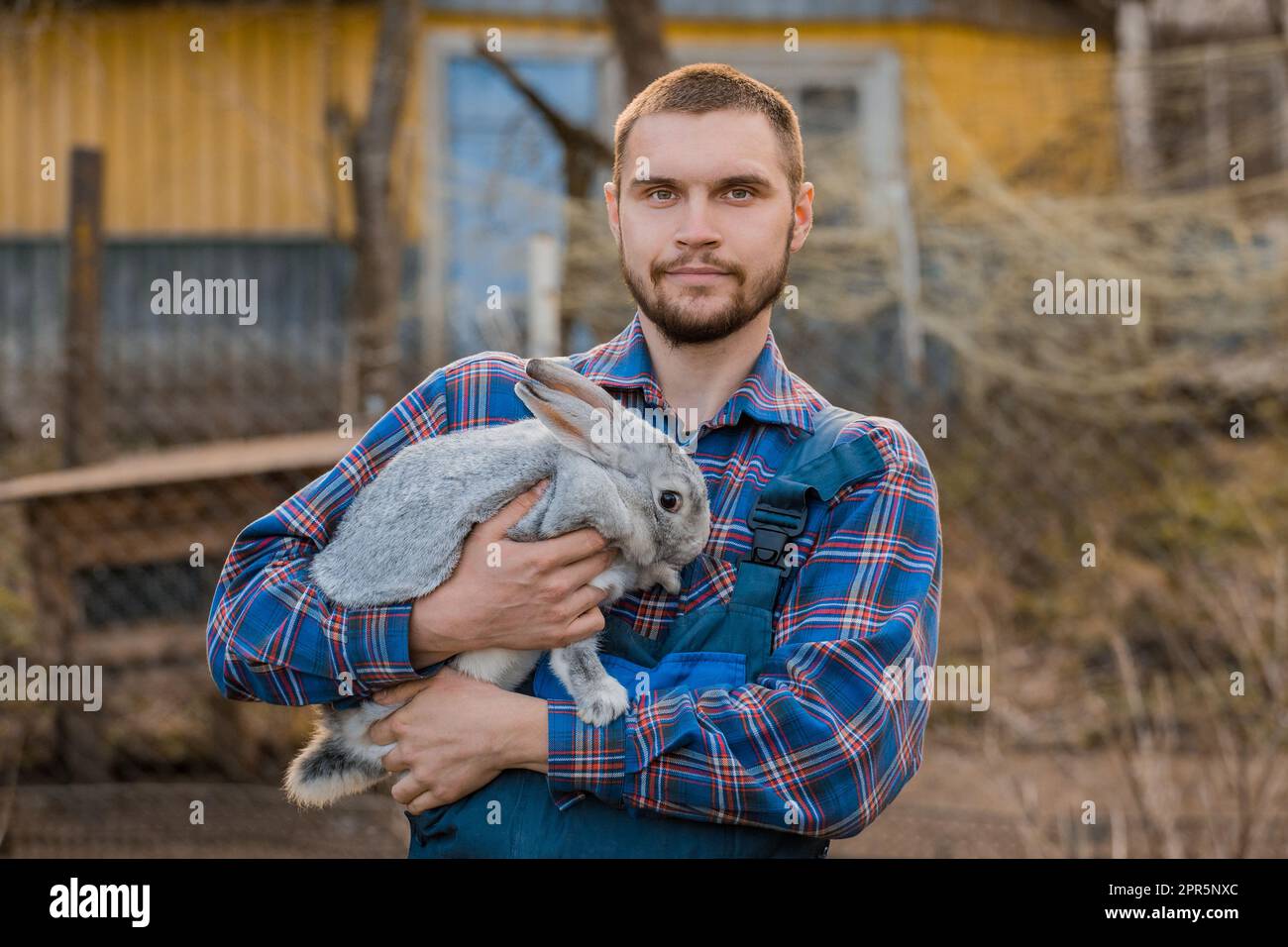 Contadino bello soddisfatto ritratto rurale caucasico europeo in campagna con barba, camicia e tute guardando la macchina fotografica con coniglio bianco nella sua A. Foto Stock