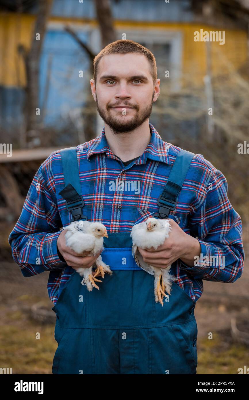 Un uomo contadino caucasico soddisfatto ritratto sorridente in una camicia e tute, tiene due nani polli bianchi in primo piano nelle sue mani pollame agricolo Foto Stock