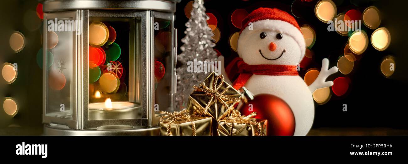 Cartolina di Natale: Pupazzo di neve, lanterna con candele, scatole luminose e lucenti con regali e luci sfocate sullo sfondo. Foto Stock