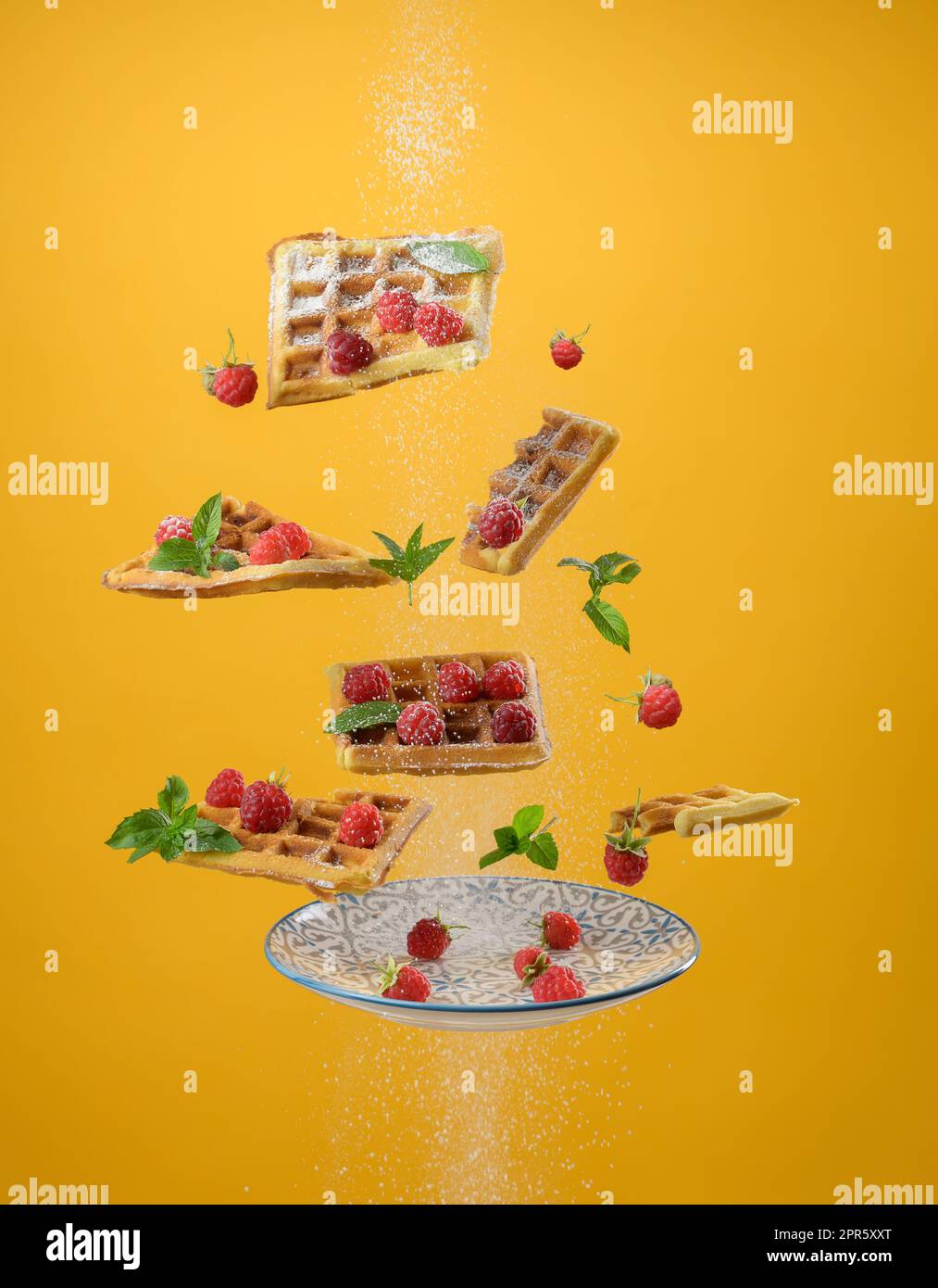 I waffle belgi al forno, i lamponi e le foglie di menta si levitano su sfondo giallo. Cospargere lo zucchero in polvere sulla parte superiore Foto Stock