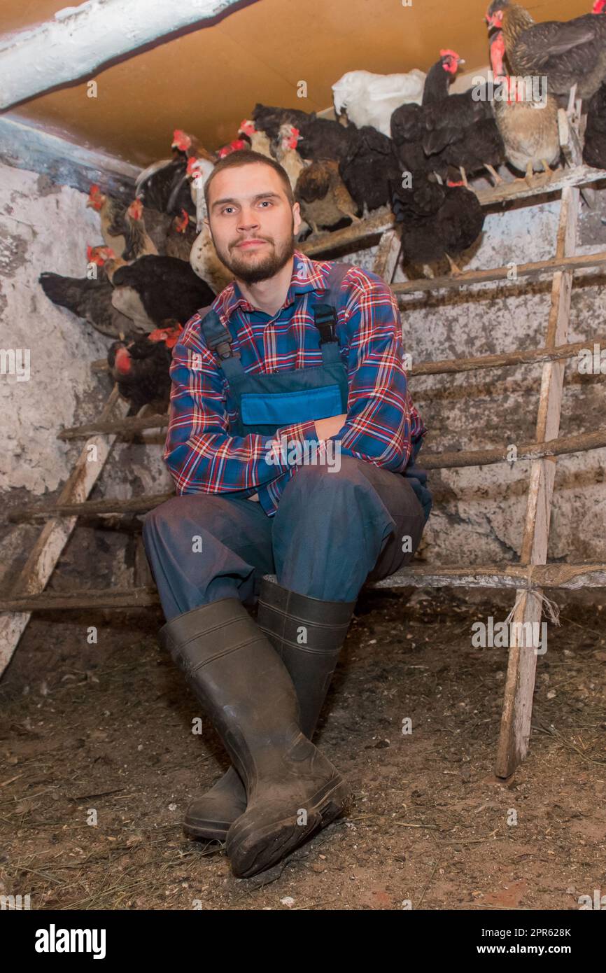 Un coltivatore soddisfatto di aspetto caucasico, giovane, bello, con una barba in camicia e tute accanto ad un persico per polli. Pollame e famiglia. Foto Stock