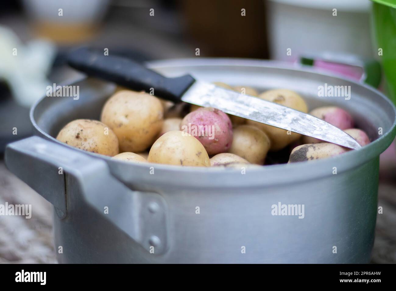 Pentola in acciaio inox con patate novelle crude e un coltello da cucina. Patate non sbucciate. Primo piano di patate con pelle gialla e rossa. Pelare le patate in cucina su un tavolo rustico di legno. Foto Stock