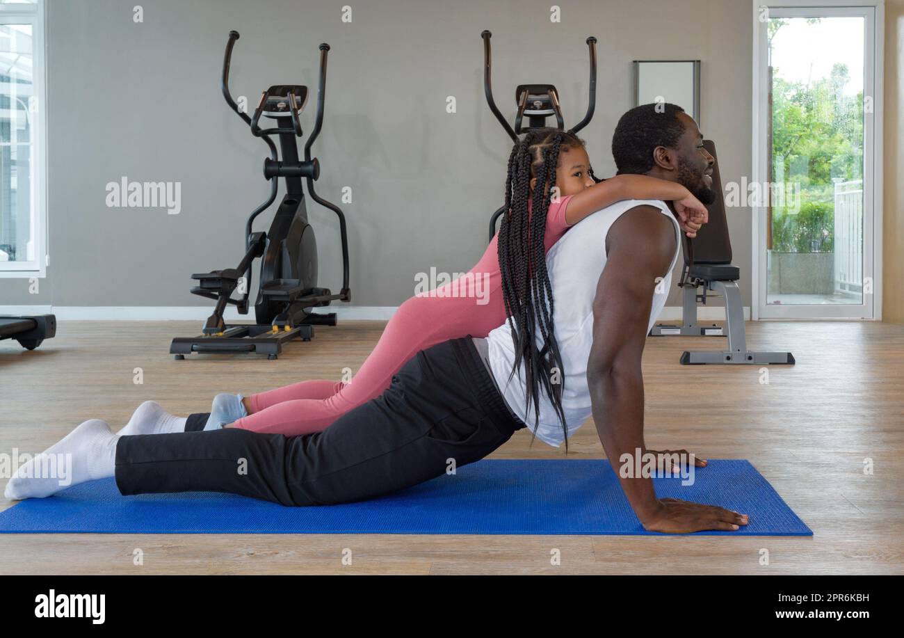 Un giovane uomo con capelli neri ricci che spinge verso l'alto il tappetino yoga mentre la figlia è sulla schiena. Felice famiglia, godetevi una vacanza insieme nel centro fitness. Foto Stock