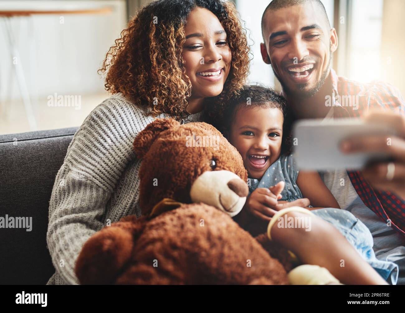 Sul conto di tre, tutti sorridono. Scatto di una bambina adorabile che prende selfie con i suoi genitori a casa su un telefono cellulare. Foto Stock