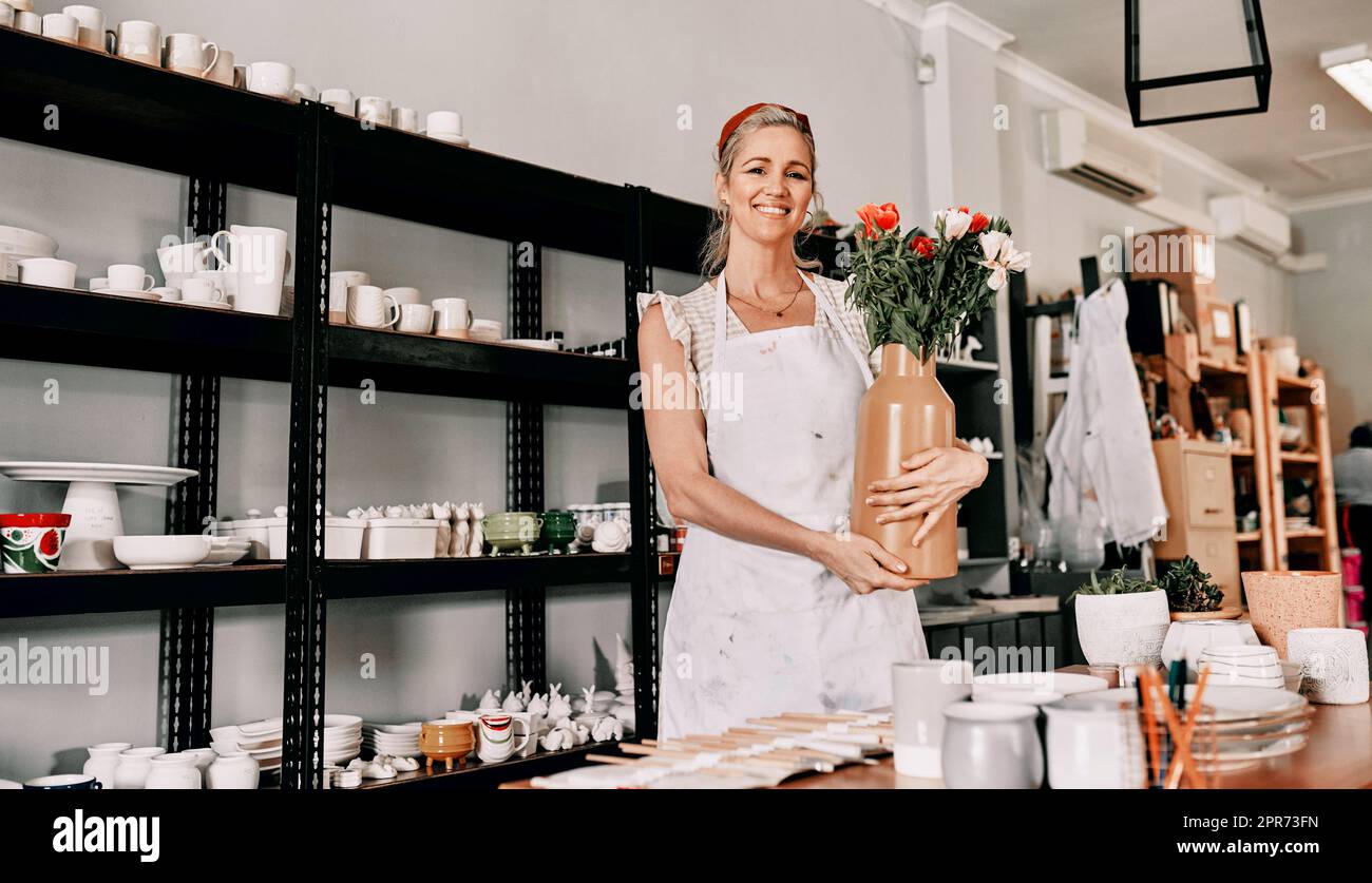 Vendiamo anche vasi. Ritratto corto di una donna matura attraente in piedi da sola e tenendo un vaso fatto a mano nel suo laboratorio di ceramica. Foto Stock