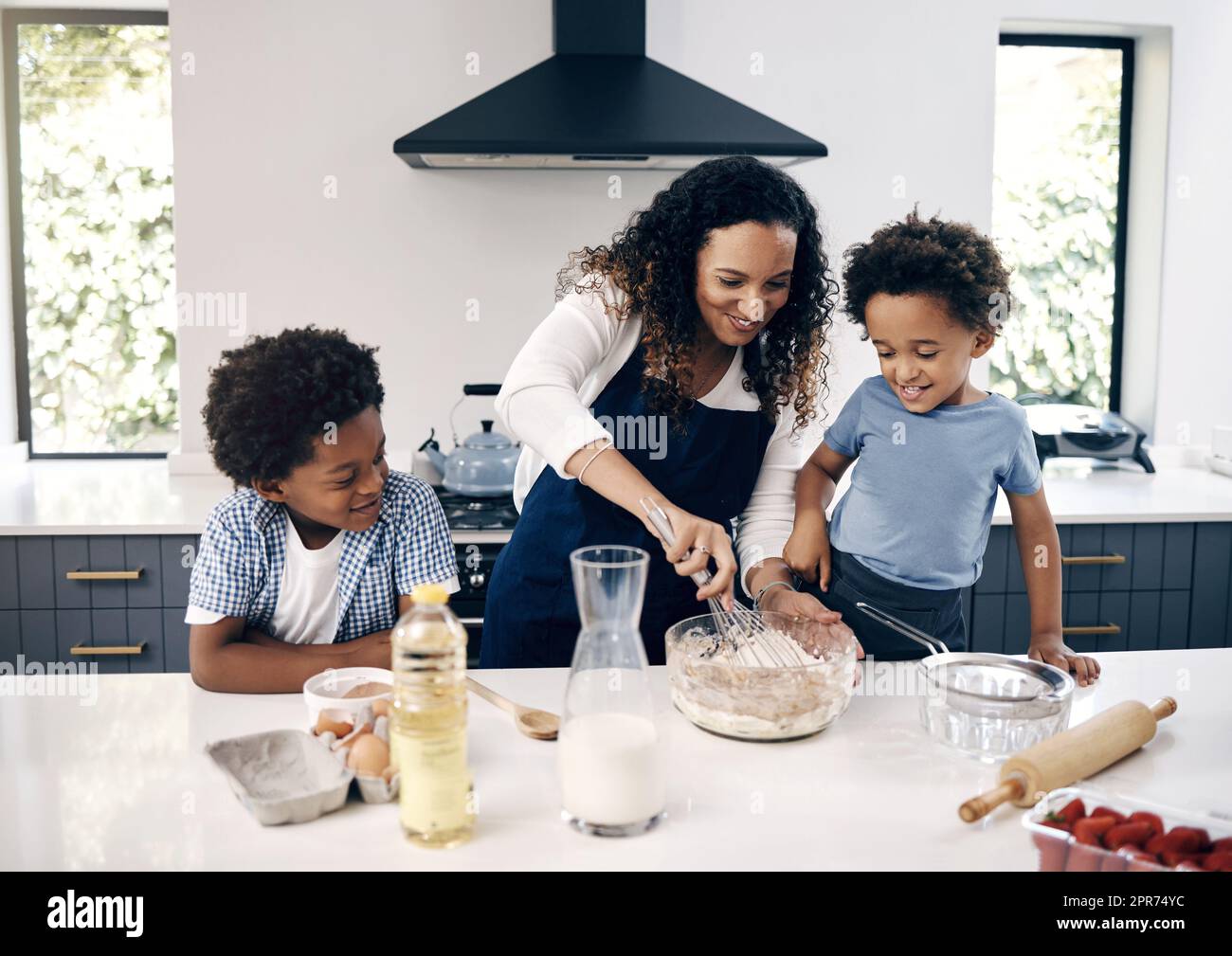 Adorabile ragazzino con afro che prepara in cucina a casa con sua madre e suo fratello. Allegra donna di razza mista che mescola ingredienti con l'aiuto dei suoi bambini. La cottura è un'attività di incollaggio Foto Stock
