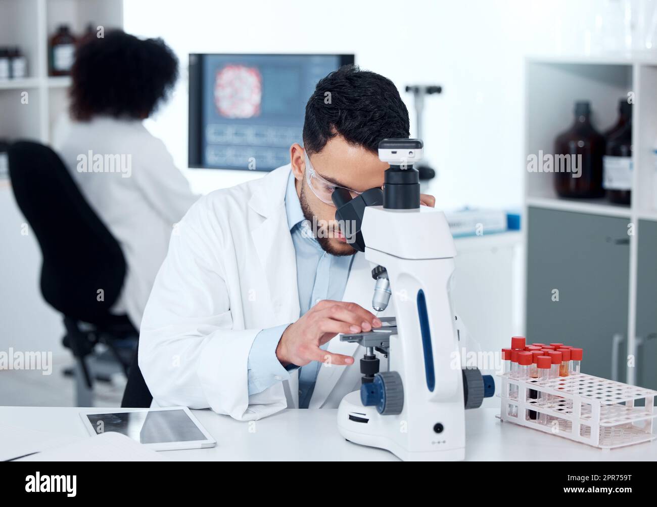 Un serio scienziato medico seduto a una scrivania e che utilizza un microscopio per esaminare e analizzare i campioni di test su vetrini. Professionista sanitario ispanico che scopre una cura per le malattie nel suo laboratorio Foto Stock