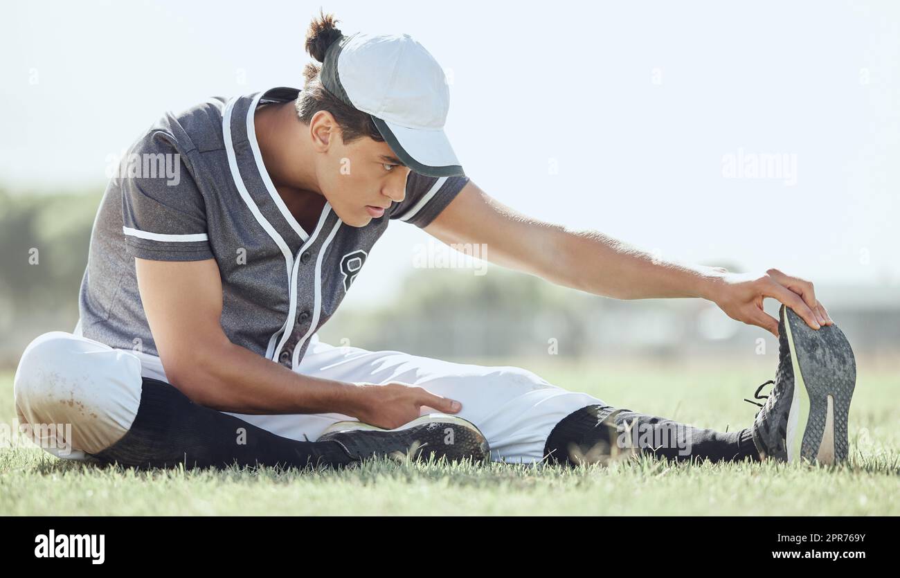 Lunghezza completa di un giocatore di baseball seduto su un campo e che si allunga prima di giocare a una partita. Atleta serio e concentrato che si prepara a giocare su erba. Uomo in forma, attivo, sportivo e atletico Foto Stock