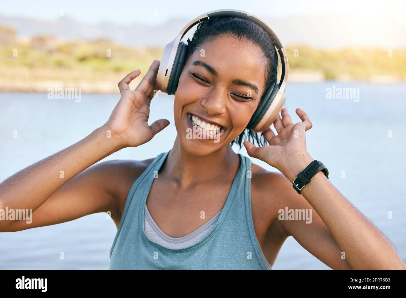 Donna eccitata che sorride e ascolta musica con l'acqua in sottofondo. Felice giovane donna che ascolta i brani della sua playlist in streaming online mentre si trova all'aperto. Utilizzando le cuffie wireless, è possibile riprodurre brani musicali Foto Stock