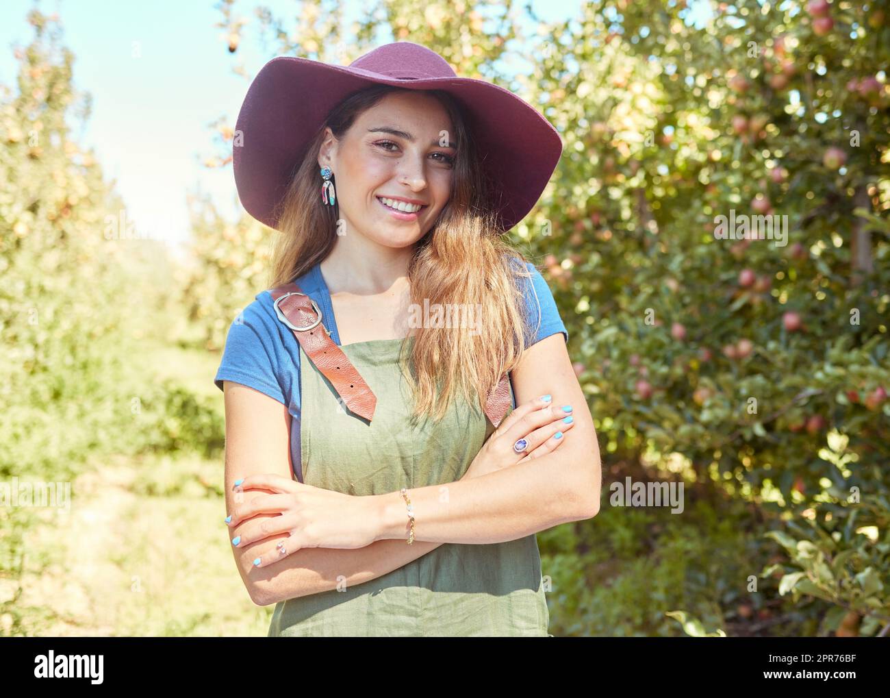 Una bella lavoratrice agricola in piedi con le braccia incrociate in una fattoria di frutta durante la stagione del raccolto. Giovane contadino felice tra alberi da frutto in estate. Produzione in crescita nell'industria agricola Foto Stock