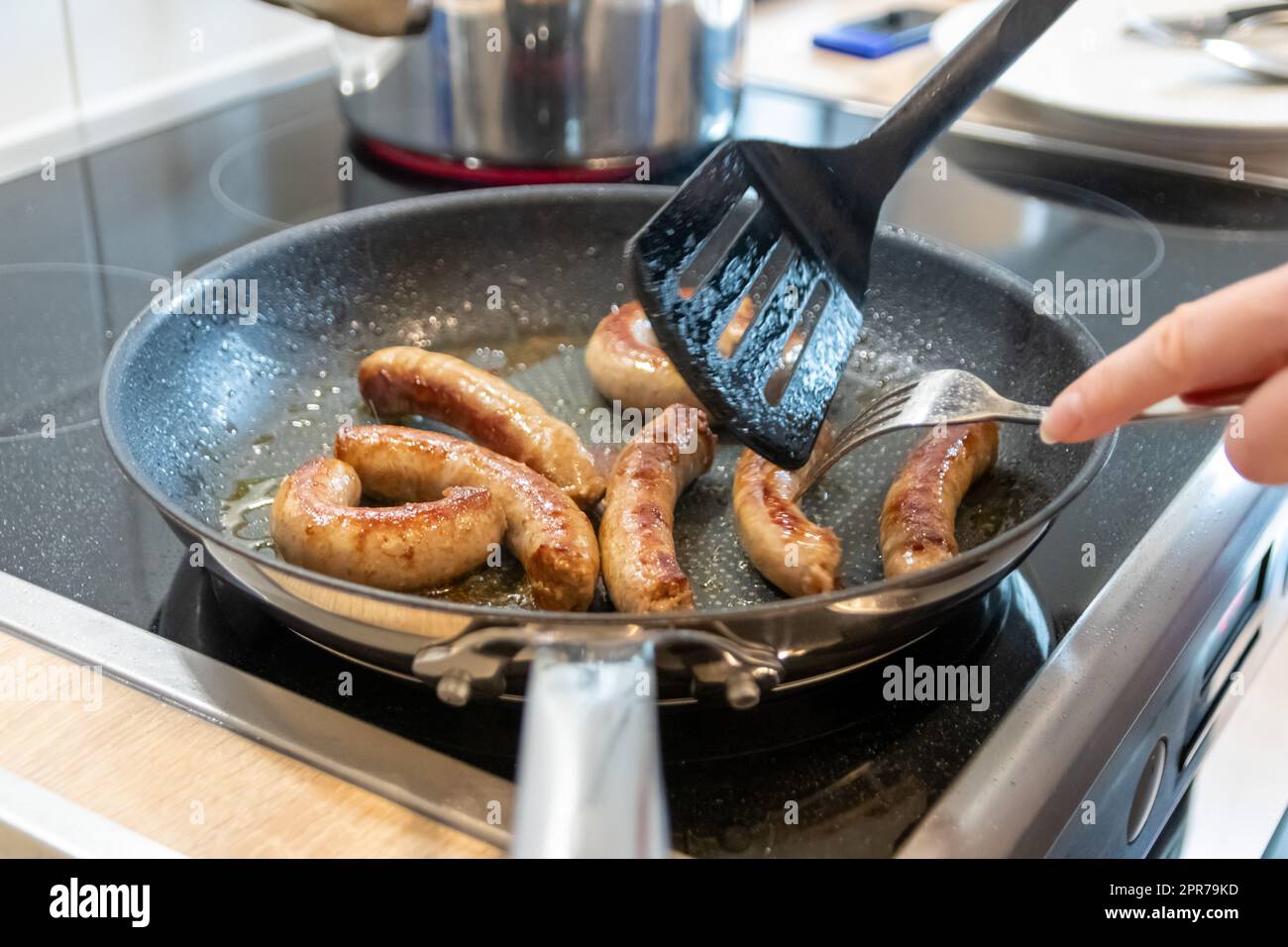 Donna che cucina salsicce in padella su piatto da cucina con forchetta e spatola nera con delizioso bratwurst grasso per cena come pasto malsano ma piccante e arrosto di salsiccia per pranzo e i bambini Foto Stock