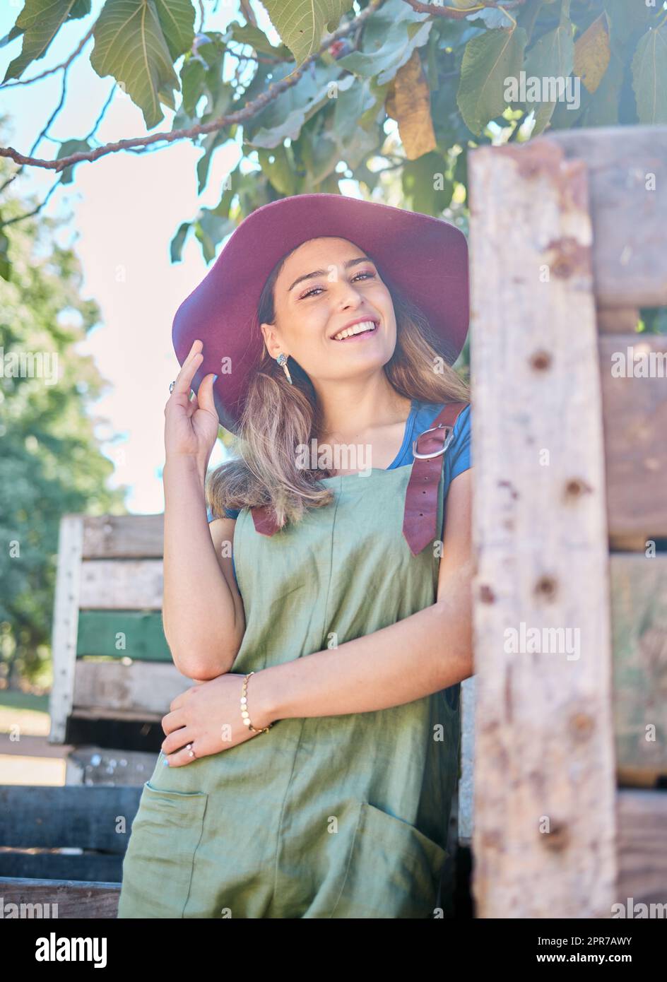 Ritratto di una donna in un cappello di paglia in piedi sotto un albero accanto a una cassa rustica di legno. Una giovane donna felice che indossa un cappello estivo e un abito dungaree in giardino in una giornata di sole a raccogliere mele Foto Stock