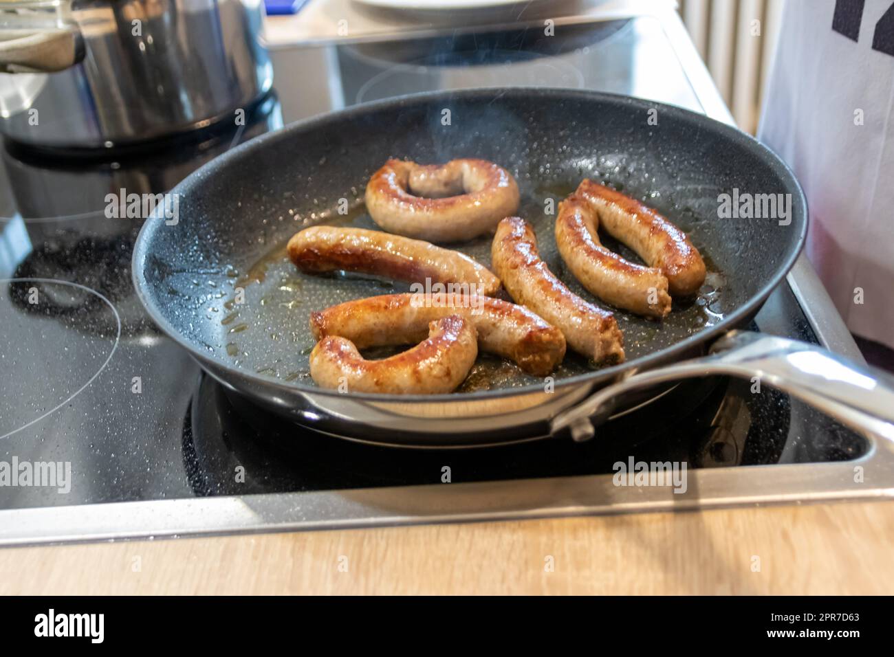Donna che cucina salsicce in padella su piatto da cucina con forchetta e spatola nera con delizioso bratwurst grasso per cena come pasto malsano ma piccante e arrosto di salsiccia per pranzo e i bambini Foto Stock