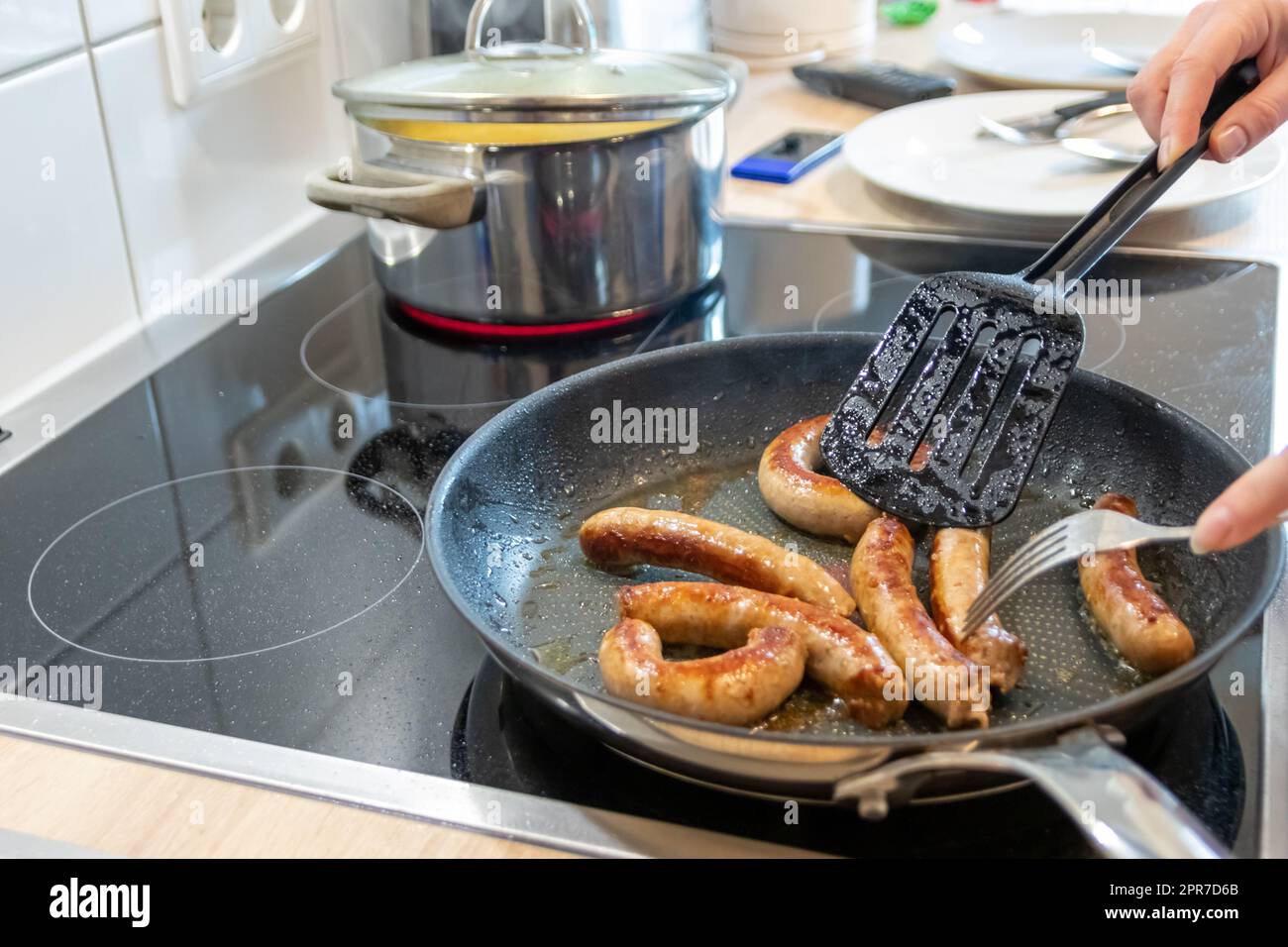 Donna che cucina salsicce in padella su piatto da cucina con forchetta e spatola nera con delizioso bratwurst grasso per cena come pasto malsano ma piccante e arrosto di salsiccia per pranzo e i bambini Foto Stock