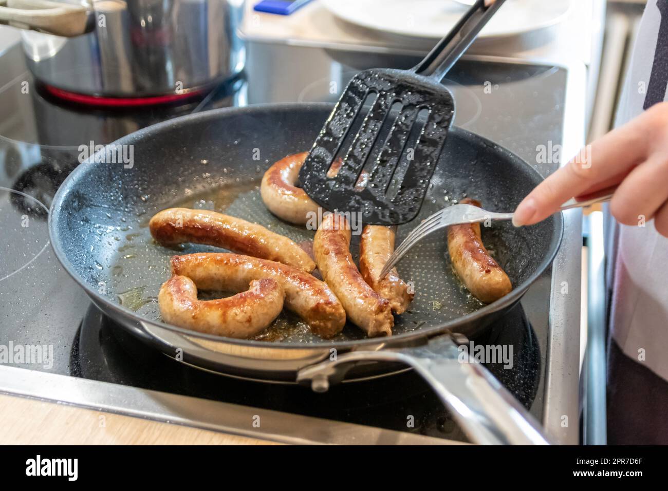Donna che cucina salsicce in padella su piatto da cucina con forchetta e spatola nera con delizioso bratwurst grasso per cena come pasto malsano ma piccante e arrosto di salsiccia per pranzo e i bambini Foto Stock
