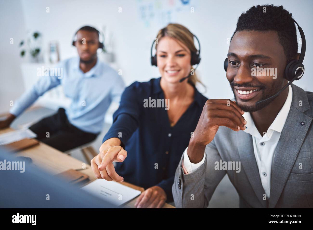 Agente di telemarketing maschile afroamericano felice che discute i piani con diversi colleghi mentre lavora insieme al computer in un ufficio. Soluzione per la risoluzione dei problemi dei consulenti per l'assistenza clienti e l'assistenza alle vendite Foto Stock