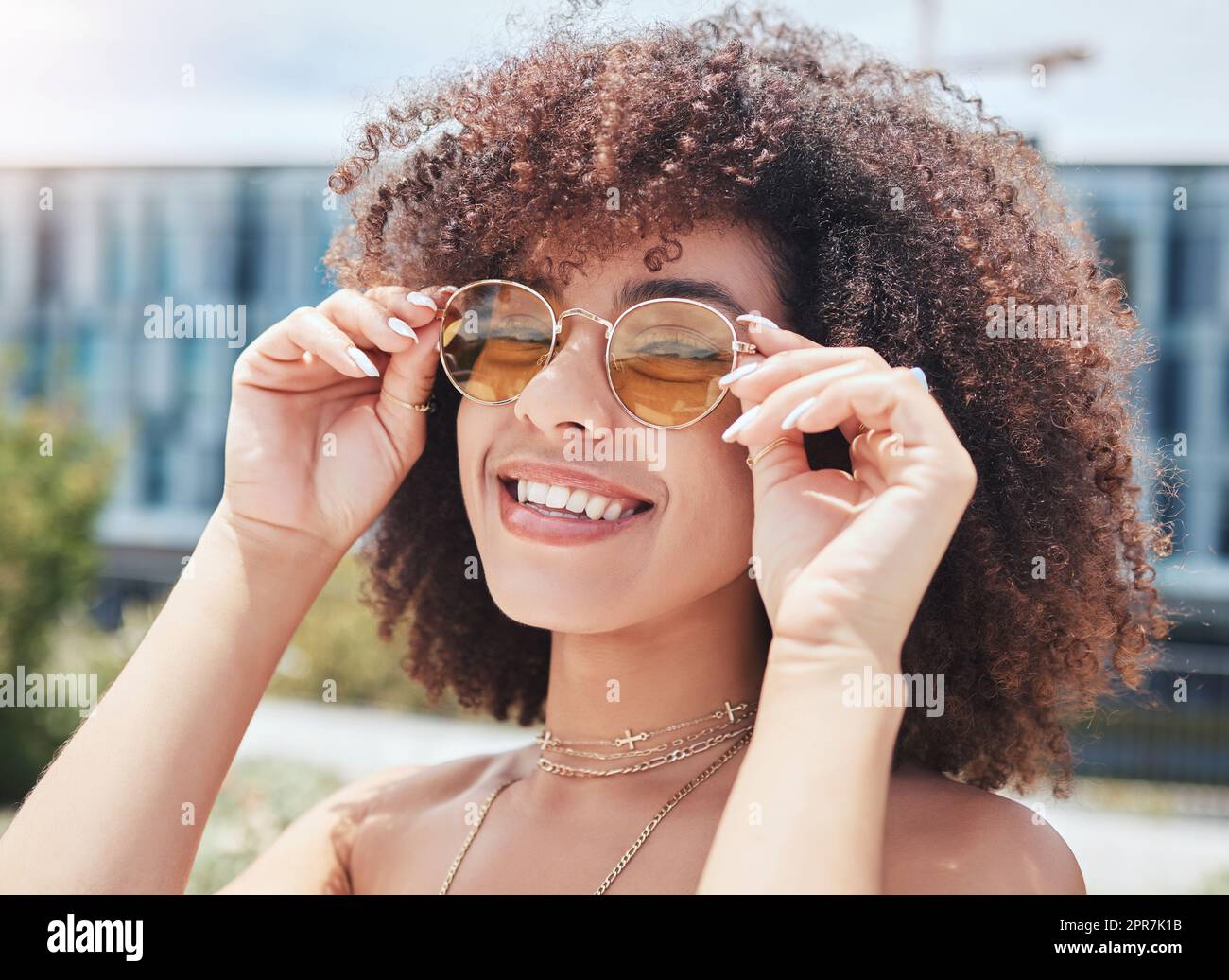 Ritratto di una giovane donna di tendenza con un afro sorridente e in posa da sola all'esterno. Donna ispanica che indossa occhiali da sole e si sente felice. Donna afroamericana alla moda in città Foto Stock