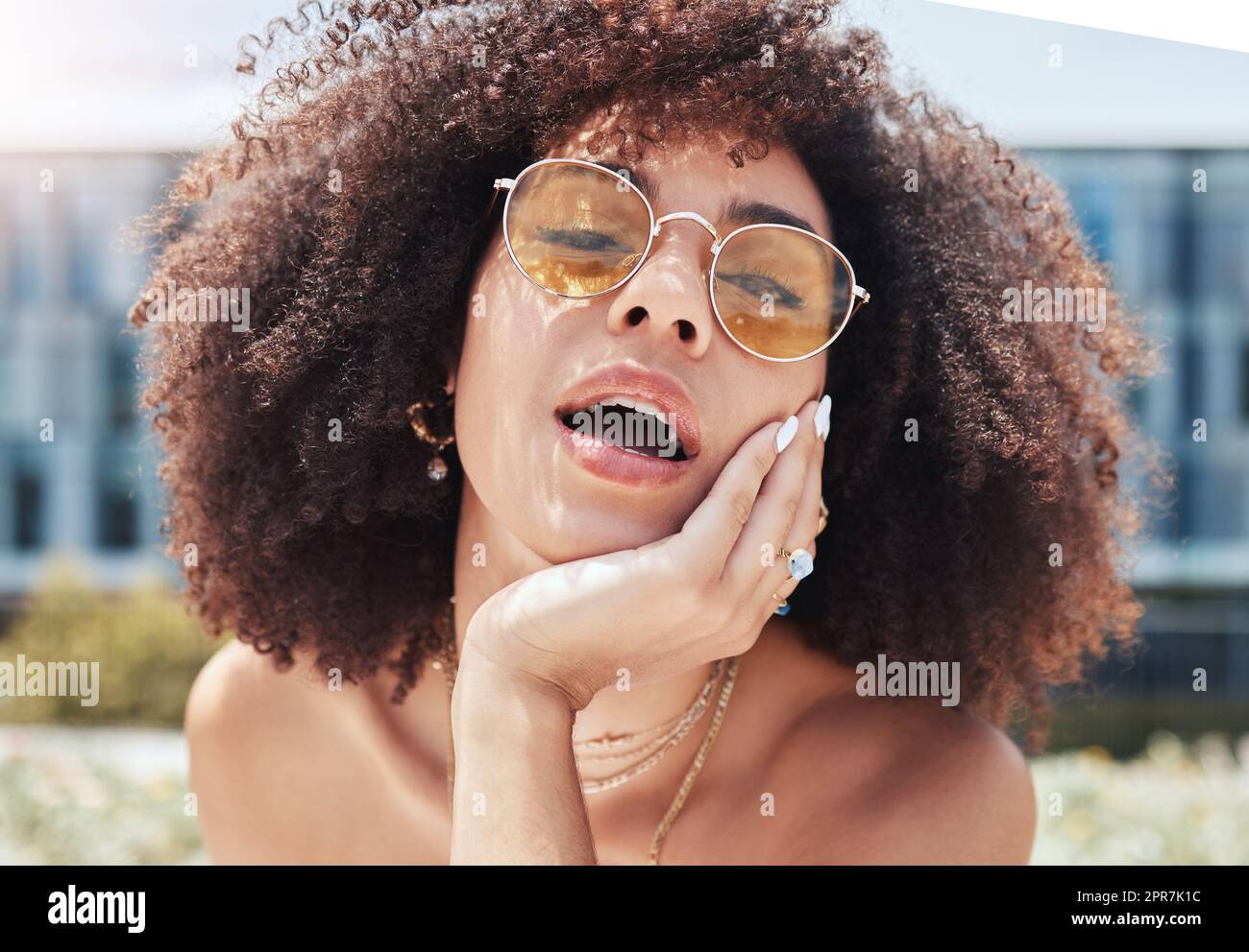 Ritratto di una giovane donna di tendenza con un afro sorridente e in posa da sola all'esterno. Donna ispanica che indossa occhiali da sole e si sente felice. Donna afroamericana alla moda in città Foto Stock