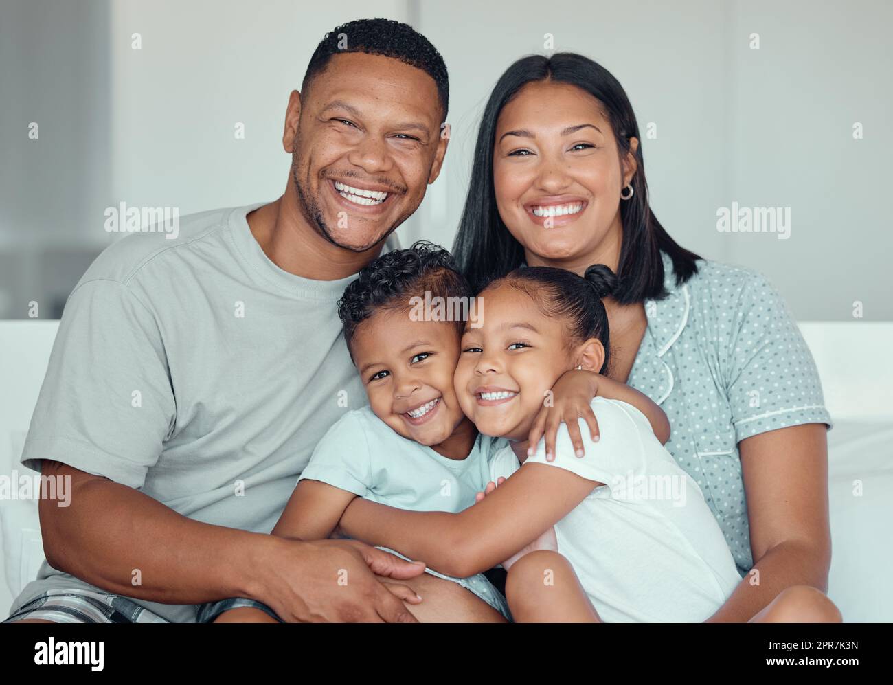 Ritratto di una giovane famiglia di razza mista con due bambini che indossano pigiama e seduti a casa insieme. Amiamo sorellina e fratello che si abbracciano mentre trascorrono del tempo con i loro genitori nel fine settimana Foto Stock