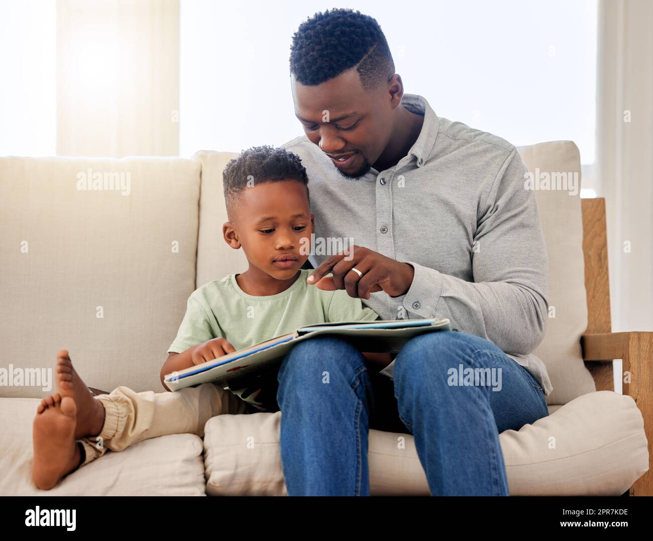 Papà afro-americano che indica la foto in un libro di favole. Adorabile bambino seduto con suo padre e leggendo un libro di favole insieme sul divano di casa. Padre e figlio passano del tempo insieme Foto Stock