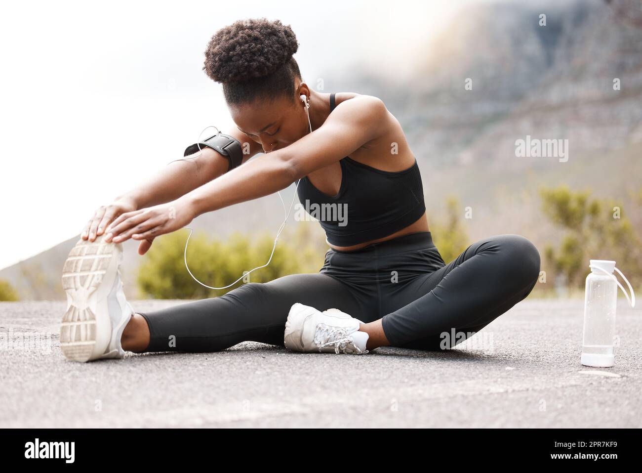 Un'atleta afroamericana con un afro che ascolta musica sulle sue cuffie mentre si esercita all'aperto in natura. Donna nera dedicata che sorride mentre si riscalda prima di un allenamento all'aperto Foto Stock