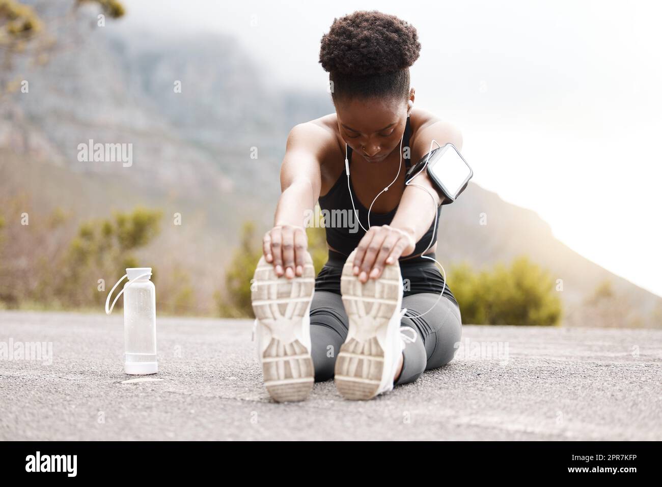 Un'atleta afroamericana con un afro che ascolta musica sulle sue cuffie mentre si esercita all'aperto in natura. Donna nera dedicata che sorride mentre si riscalda prima di un allenamento all'aperto Foto Stock