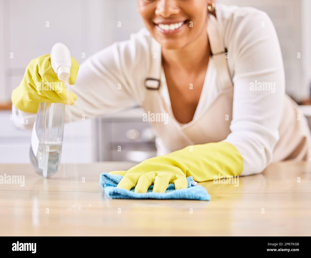 Detergente femminile felice e irriconoscibile sorridente mentre spruzza e pulisce una superficie del tavolo con un panno che pulisce da solo a casa. Una donna che pulisce un bancone mentre indossa i guanti Foto Stock