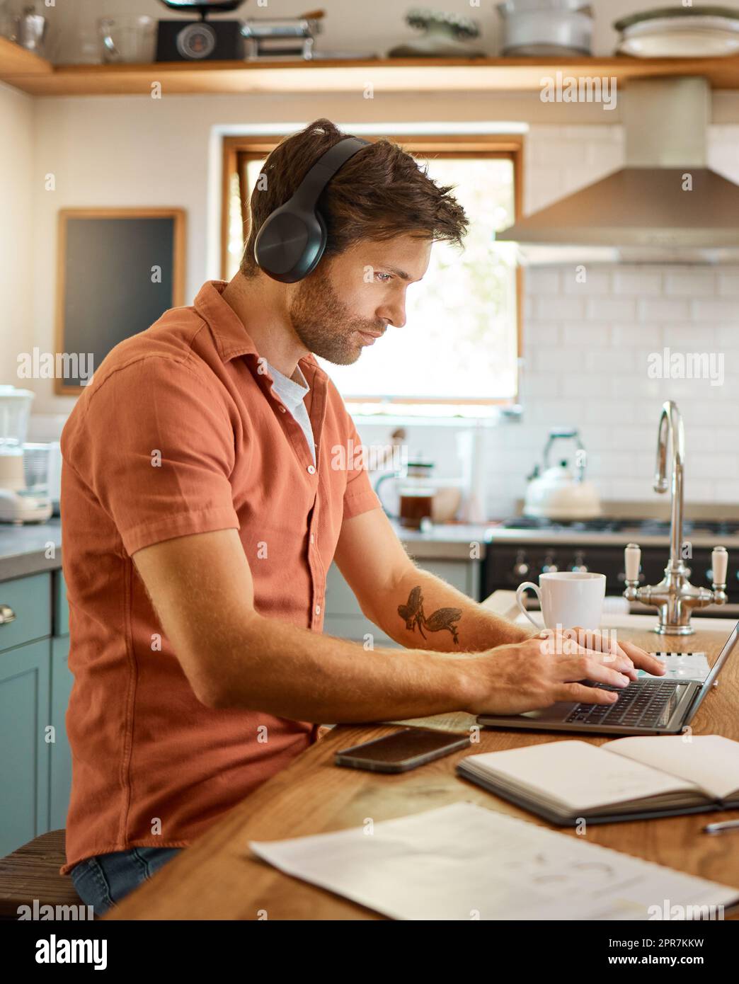 Giovane uomo d'affari caucasico serio che indossa cuffie e ascolta musica mentre usa un notebook da solo a casa. Un uomo d'affari maschio che digita un notebook mentre lavora in cucina a casa Foto Stock