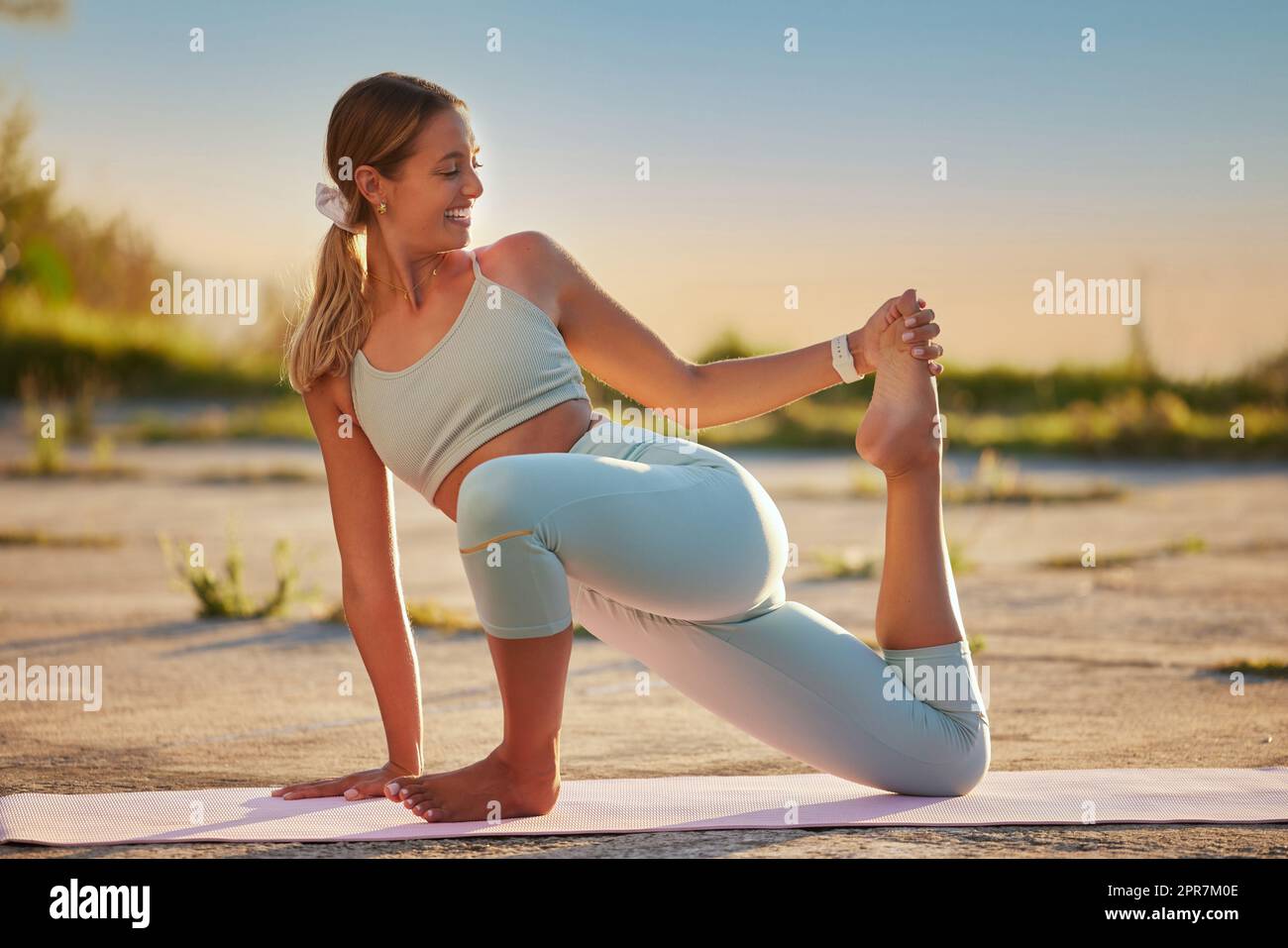 Donna yoga a lunghezza intera che tiene una lucertola ritorta in posa in pratica all'aperto in una natura remota. Sorridente bella persona caucasica che usa il tappetino, bilanciata mentre si allunga al tramonto. Giovane, attivo, zen, sereno Foto Stock