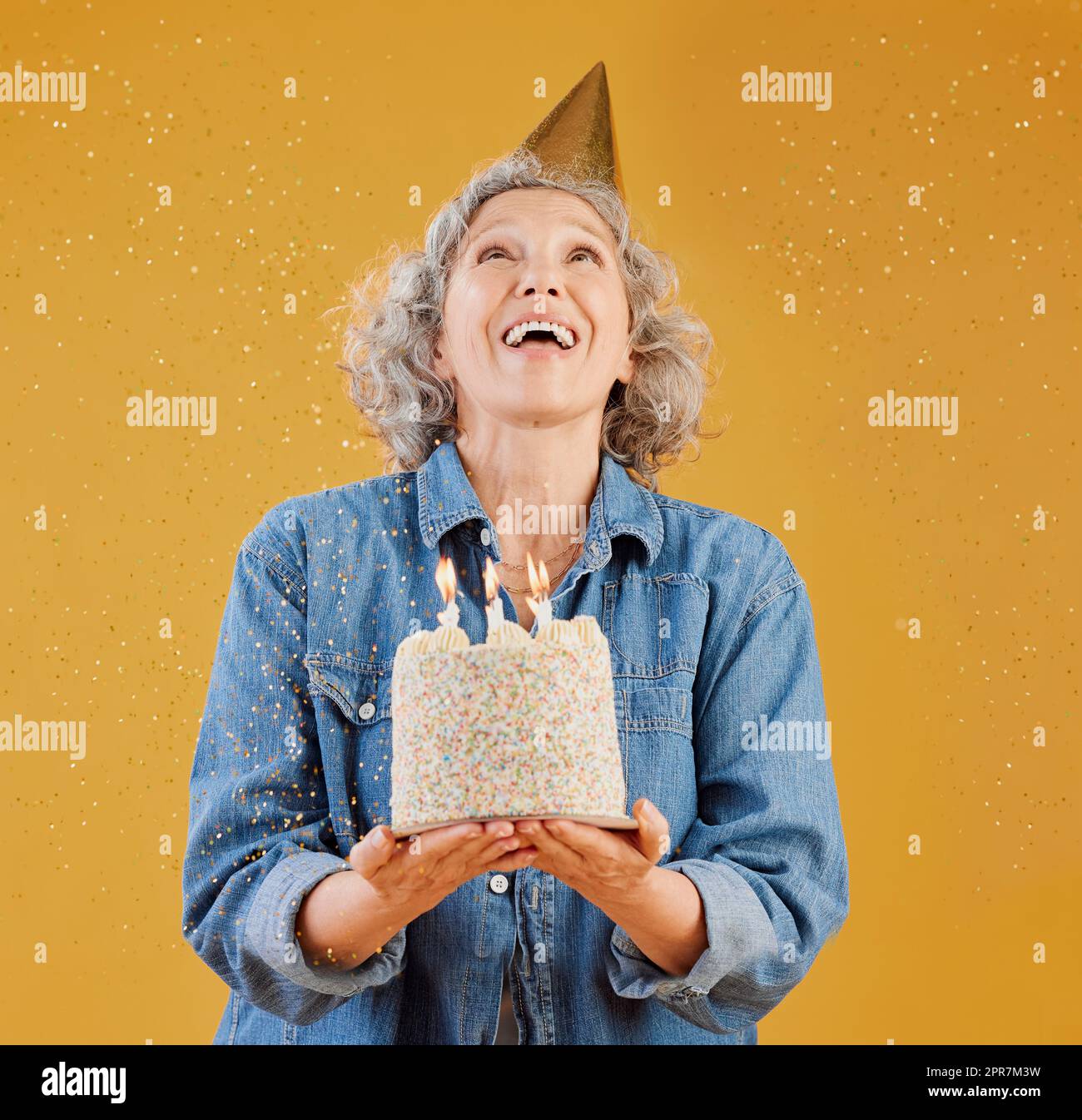 Una donna caucasica matura felice che indossa un cappello di compleanno e tiene in mano una torta mentre i coriandoli cadono dall'alto su uno sfondo giallo nello studio. Donna bianca sorridente che festeggia un altro anno Foto Stock
