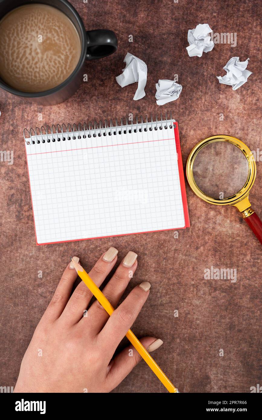 Mano di donna con notebook a spirale con spazio per la copia, matita, lente d'ingrandimento, tazza da caffè e carte schiacciate. Donna d'affari che scrive un messaggio importante su Book. Foto Stock