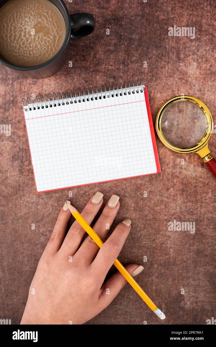 Mano di donna con notebook a spirale con spazio per la copia, matita, lente d'ingrandimento e tazza di caffè sopra il tavolo. Donna d'affari che scrive un messaggio importante su Book with drink. Foto Stock