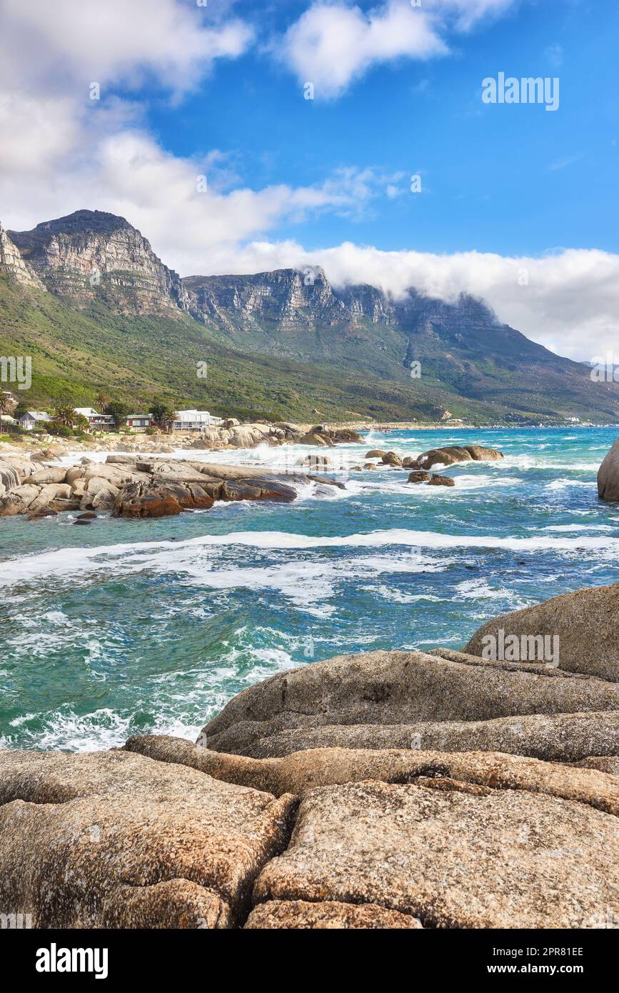 Vista panoramica della spiaggia dell'oceano, del mare, delle nuvole, del cielo blu con spazio fotocopie su Camps Bay, città del Capo, Sud Africa. Onde mareali che si gettano su rocce o massi della costa. Sfondo dei dodici monti Apostoli Foto Stock